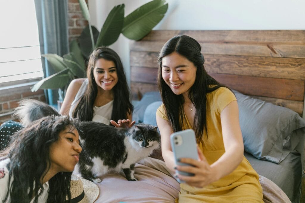 Three women smiling and taking a selfie with a cat in a cozy bedroom setting.
