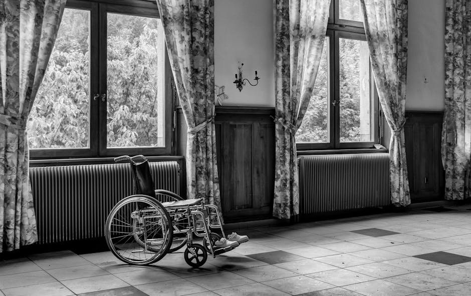 Black and white photo of an empty room with a wheelchair by large windows.